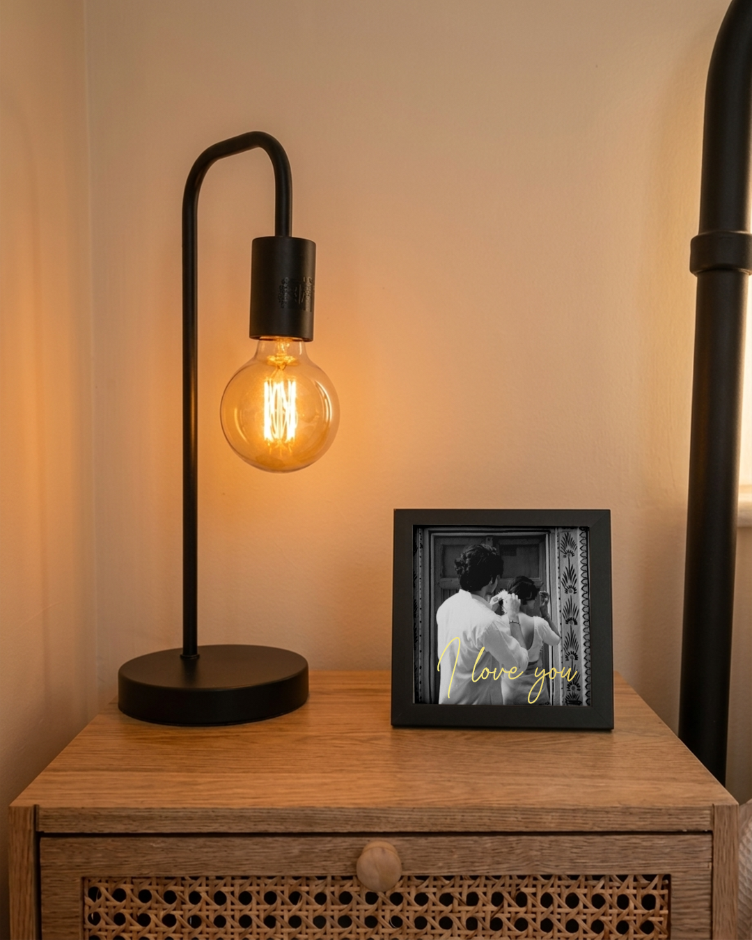Black lamp with a lit bulb on a wooden surface next to a framed photo with 'I love you' text.